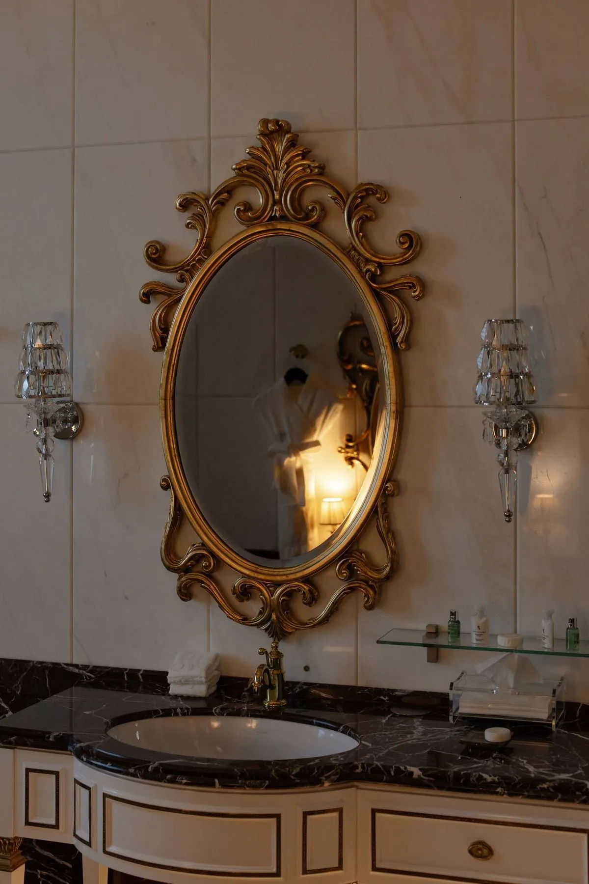 Bathroom sink with an ornate oval mirror and classic wall lamps.
