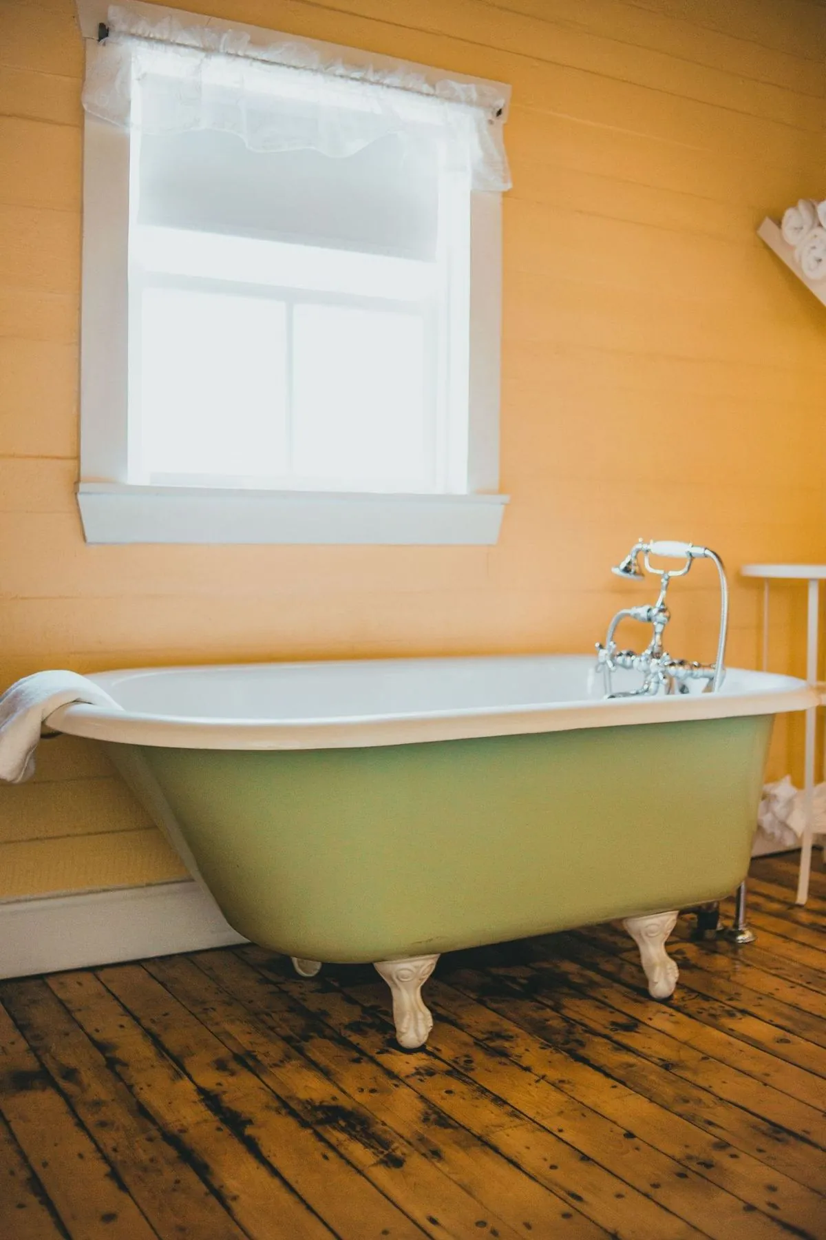 Sage-green clawfoot tub by a bright window in a rustic bathroom.